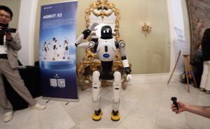 Humanoidalny robot AgiBot X2 w biało-czarnej obudowie macha ręką podczas pokazu w czasie targów MWC 2026, obok baner z nazwą modelu