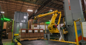 Zrobotyzowany paletyzator: żółte ramię robota FANUC układa kartony na palecie przy przenośniku rolkowym, obok stoi owijarka do folii stretch