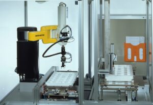 Robot SCARA z żółtym ramieniem nad tacką z detalami w stanowisku z podajnikiem tacek