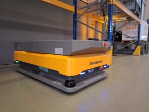 Żółto-szary robot AMR Jungheinrich arculee M z płaską platformą ładunkową na posadzce magazynu, obok regału paletowego