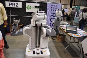 Dwuramienny robot PR2 na stoisku z banerem ROS i tabliczką TurtleBot, obok stół z laptopem i ulotkami