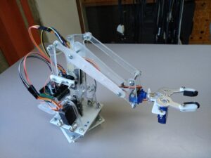 Małe ramię robota z przezroczystej pleksi, którego przeguby i chwytak poruszają niebieskie i czarne serwomechanizmy modelarskie podłączone do płytki Arduino